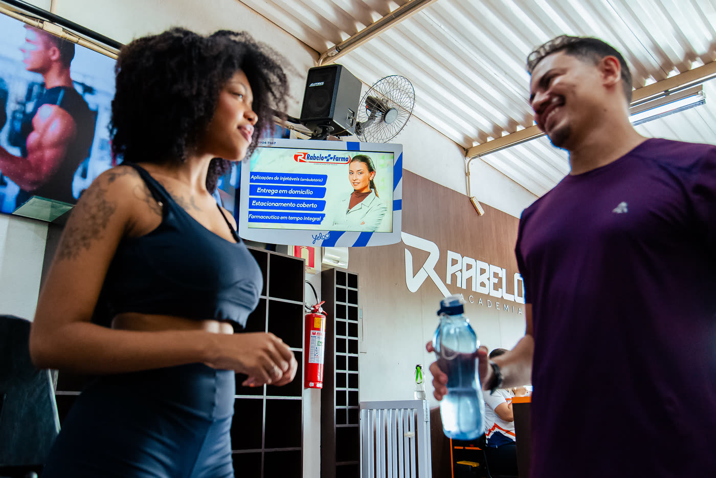 Alunos conversando na academia com uma tela de anúncio ao fundo